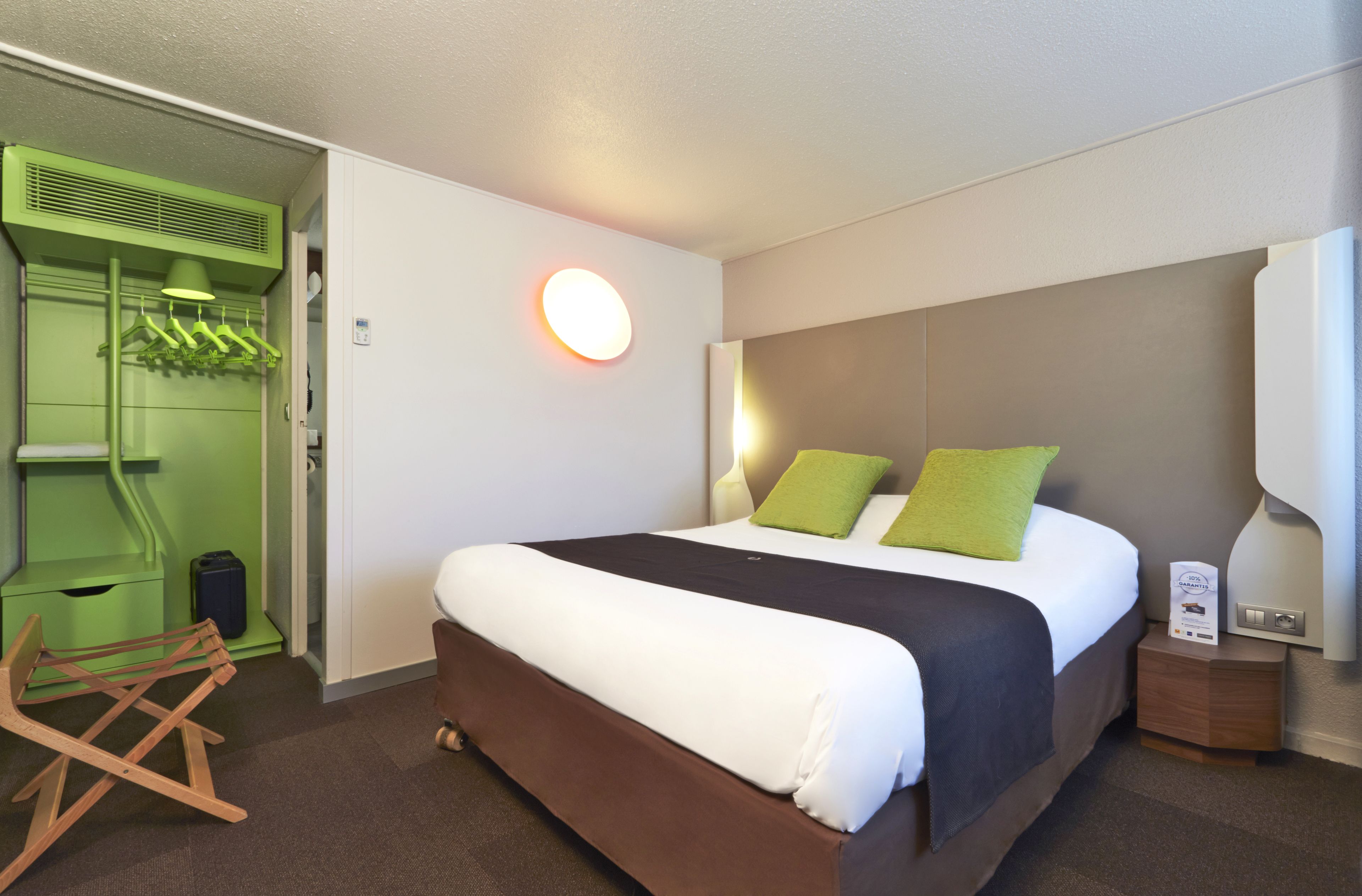 Una habitación de hotel con una cama grande, un armario verde, una silla de madera y una mesita de noche con una lámpara.