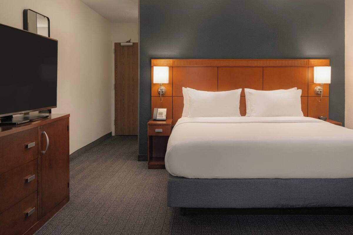 Hotelkamer met een groot bed, een houten hoofdbord, een flatscreen televisie op een houten kast en een nachtkastje met een lamp.