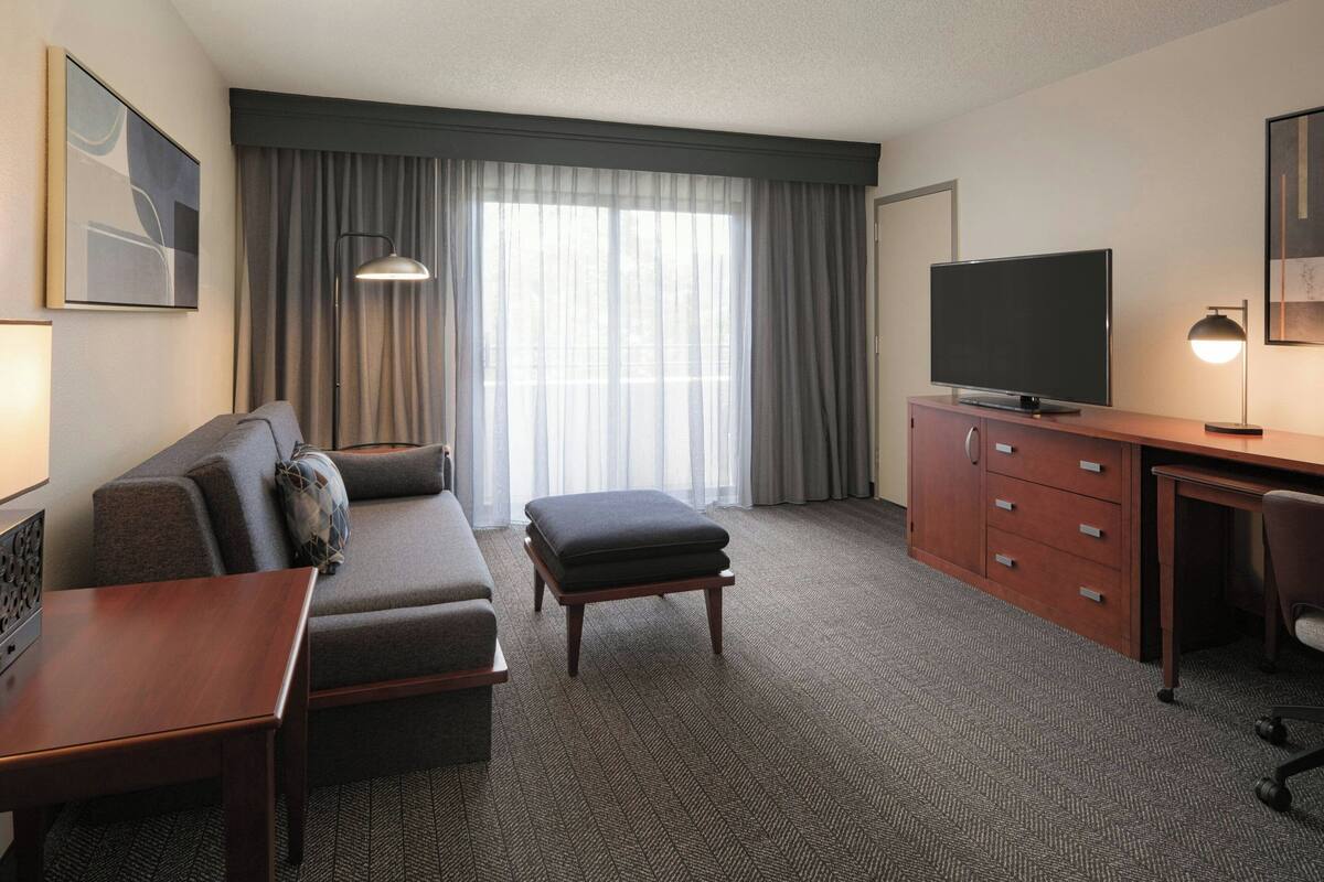 Hotelkamer met een bank, voetenbankje, bureau, televisie en een groot raam met gordijnen.