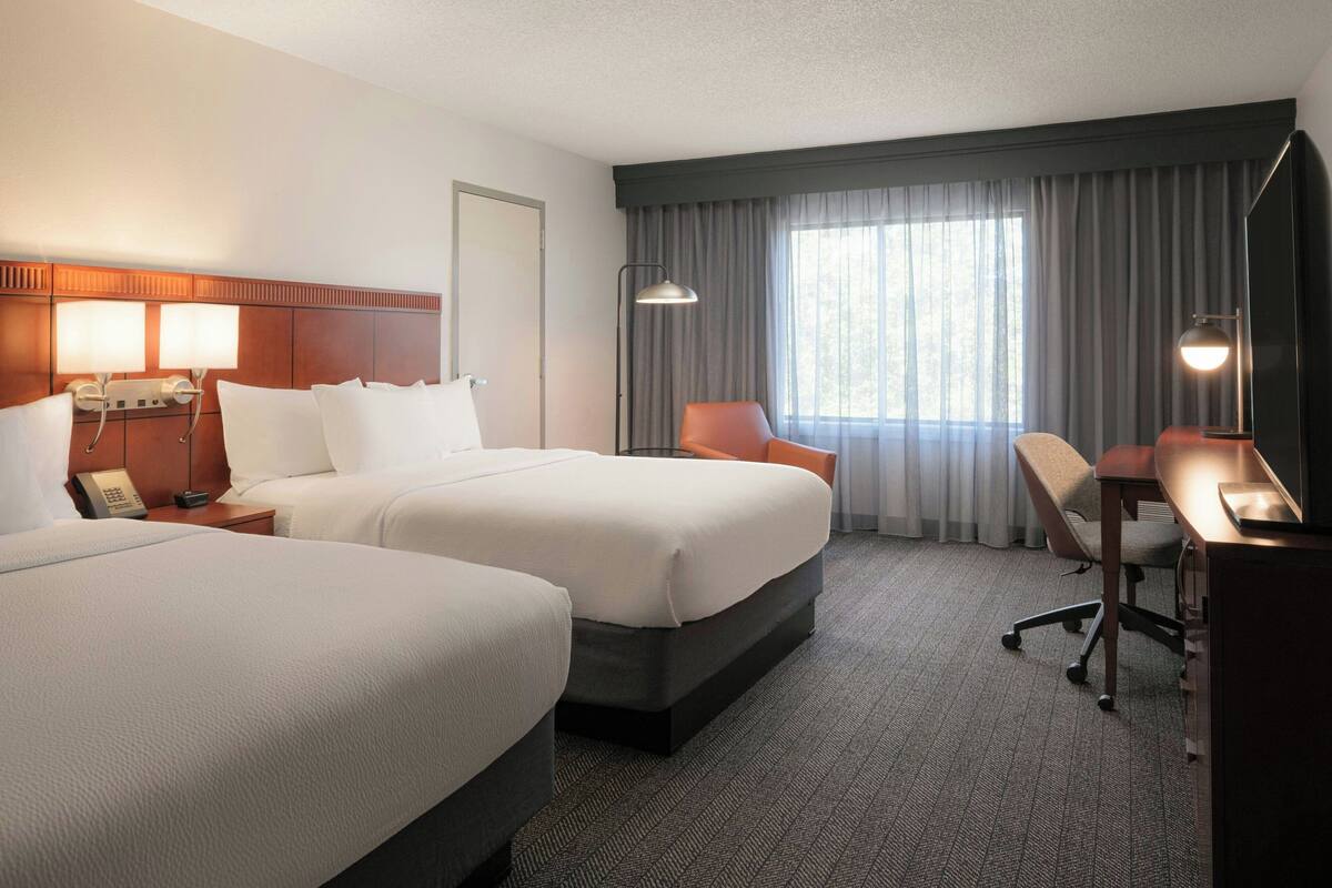 Een hotelkamer met twee bedden, een bureau, een stoel, een lamp en een raam met gordijnen.
