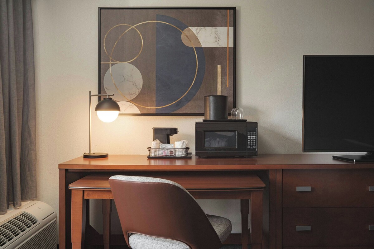 Une chambre d’hôtel comprenant un bureau en bois, une chaise, un téléviseur à écran plat, une œuvre d’art abstraite encadrée, une machine à café, un four à micro-ondes et une fenêtre avec des rideaux.