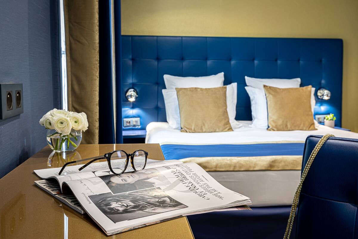 A hotel room with a blue headboard, a bed with white and brown pillows, a nightstand with a lamp, and a table with a vase of white flowers and a magazine.