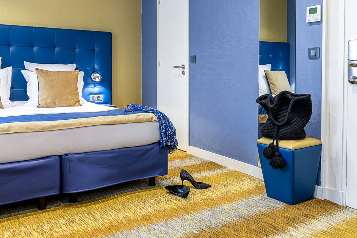 A hotel room with a large bed, a blue headboard, a small seating area with a chair and a coat rack, and a wooden floor.