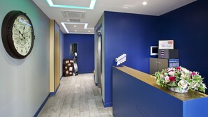 A reception area with a blue wall, a clock, a reception desk with a flower arrangement, and a digital display. - Trianon Gare de Lyon (Paris)