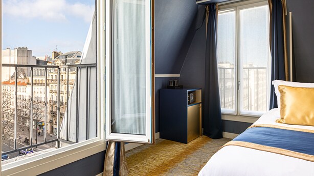 A hotel room with a large window, a bed, a desk, and a view of the city. - Trianon Gare de Lyon (Paris)