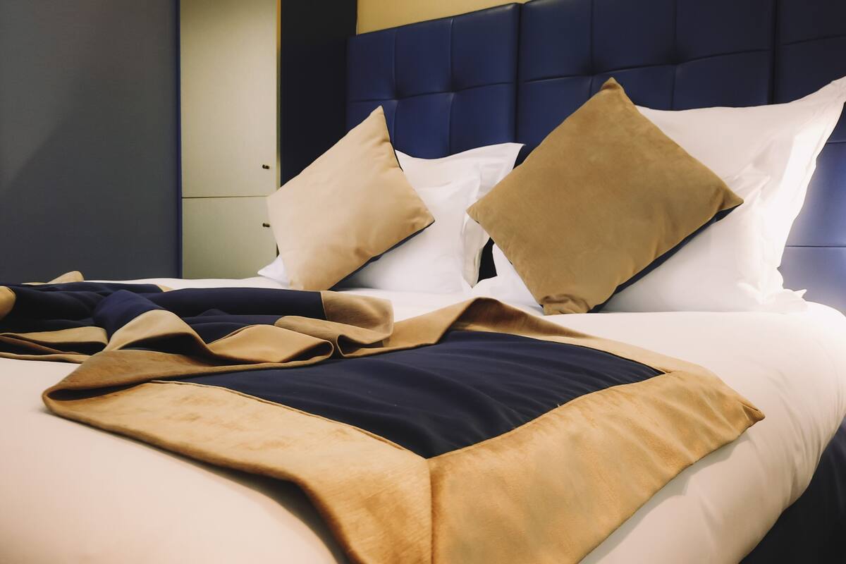 A neatly made bed with a blue headboard, multiple pillows, and a beige and navy throw blanket.