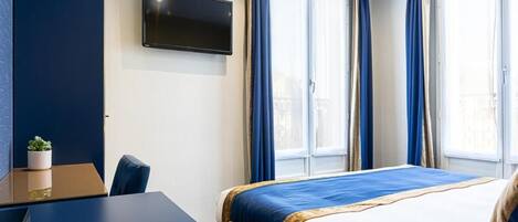 A hotel room with a bed, a desk, a TV, and large windows with curtains.
