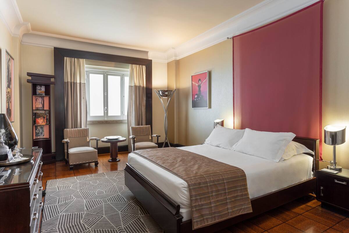 A hotel room with a large bed, a desk, a chair, a floor lamp, and a bookshelf.