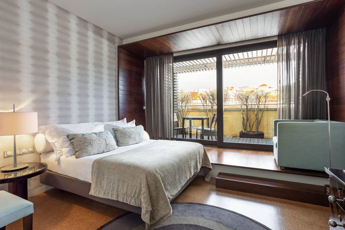 A modern hotel room with a large bed, a sitting area with a chair, a small table, and a balcony with a view of buildings and trees.