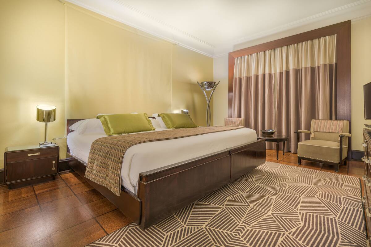 A hotel room with a large bed, a nightstand, a lamp, a chair, and a patterned rug.