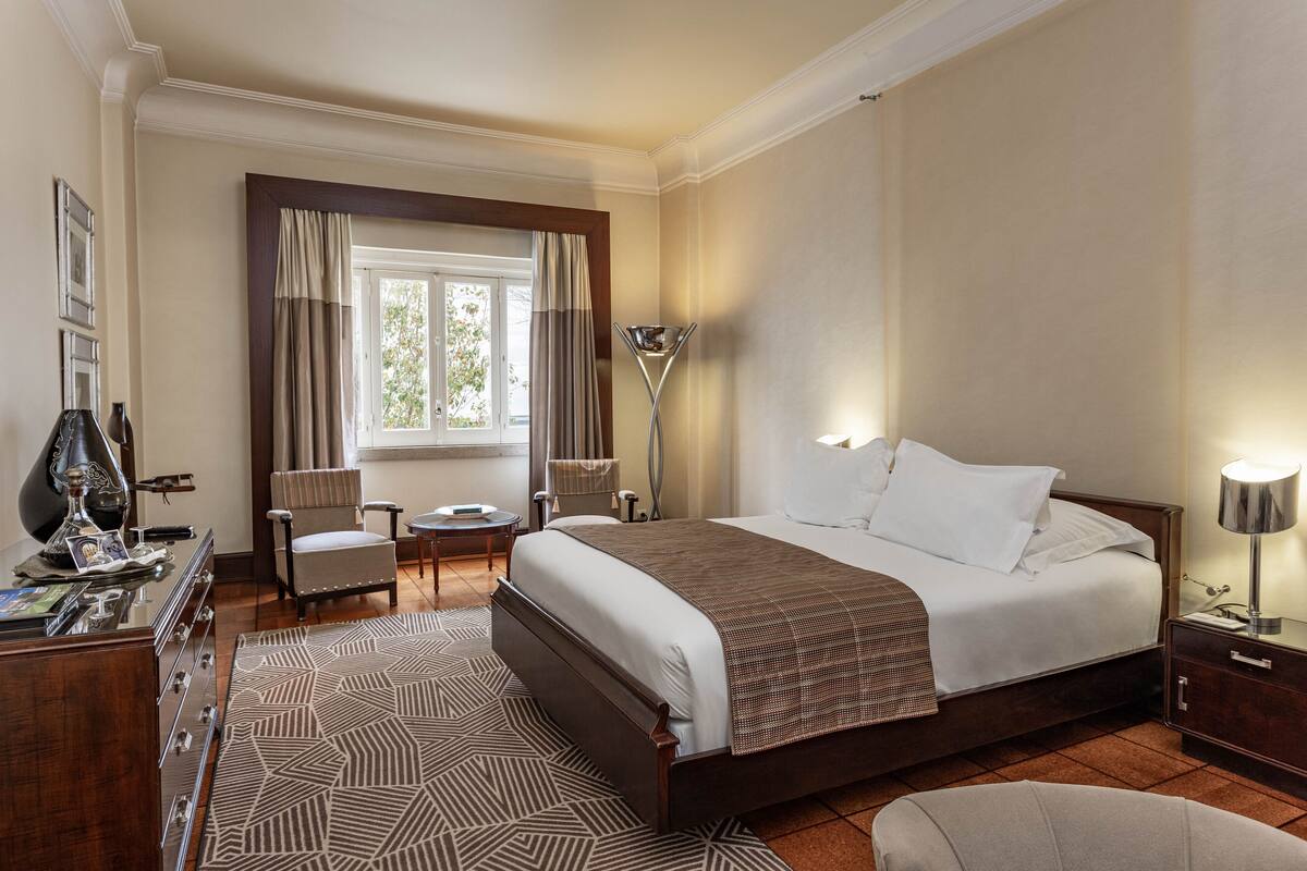 A hotel room with a bed, a desk, a chair, a lamp, and a window with curtains.