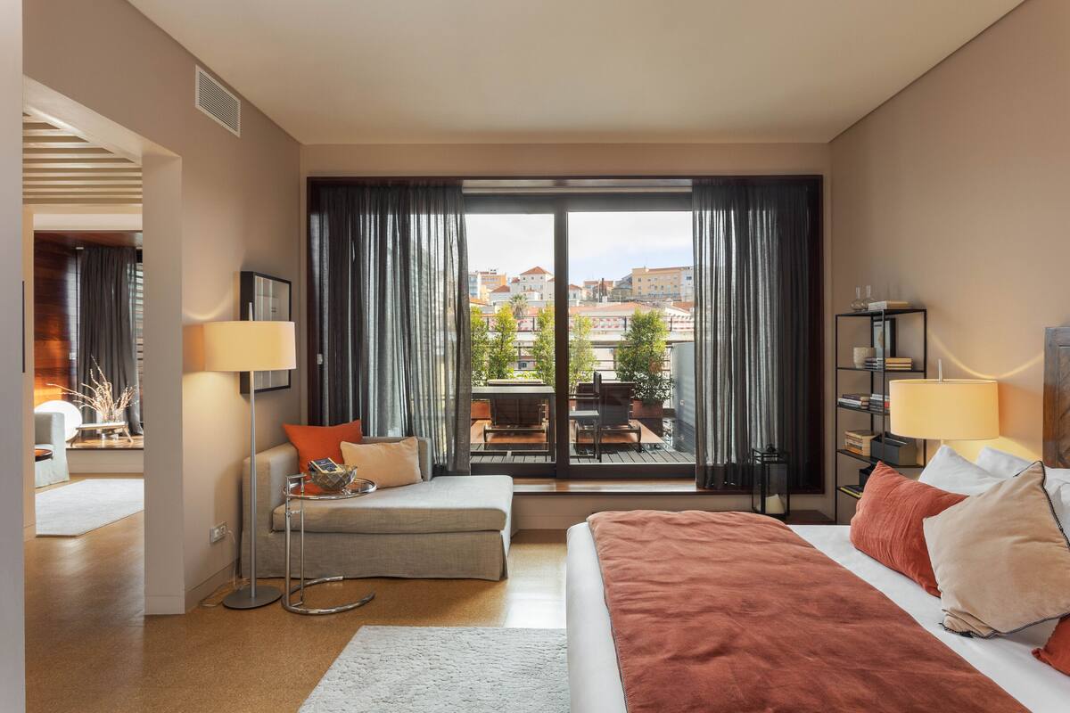 A modern hotel room with a large bed, a sofa, a desk, and a view of the city through large windows.