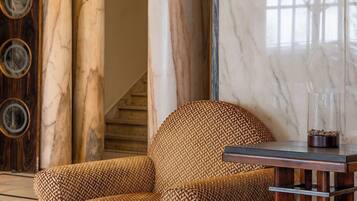 Un hall con un sillón estampado, una mesita de madera, un candelabro y columnas de mármol.