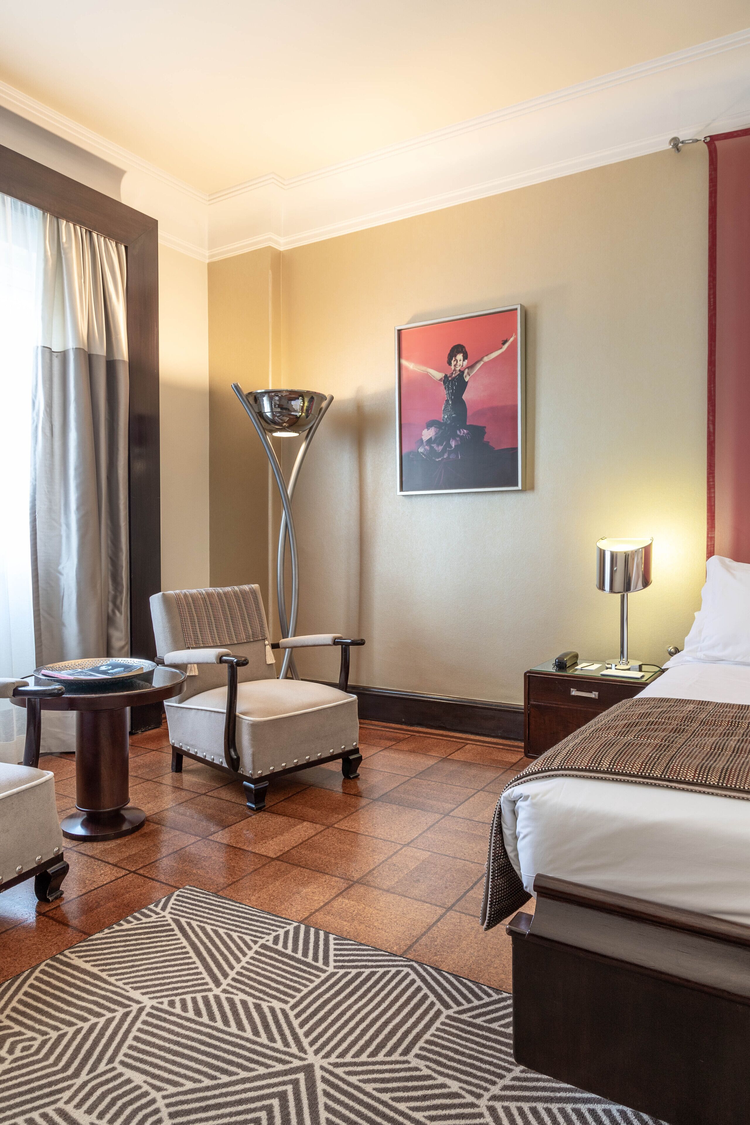 A hotel room with a bed, a chair, a lamp, a nightstand, a rug, and a painting on the wall.