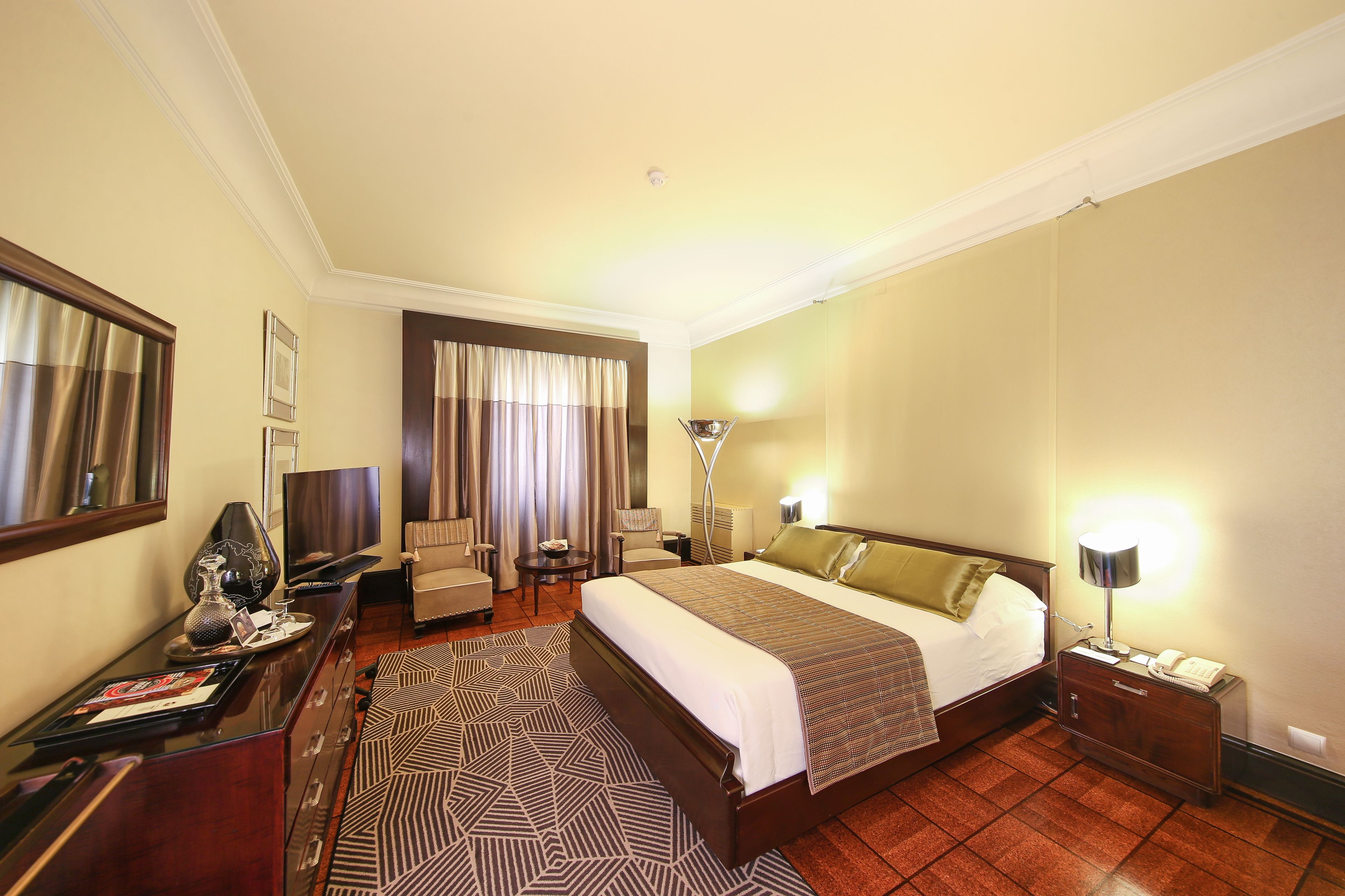 A hotel room with a large bed, a desk with a lamp, a chair, a TV, and a patterned rug.