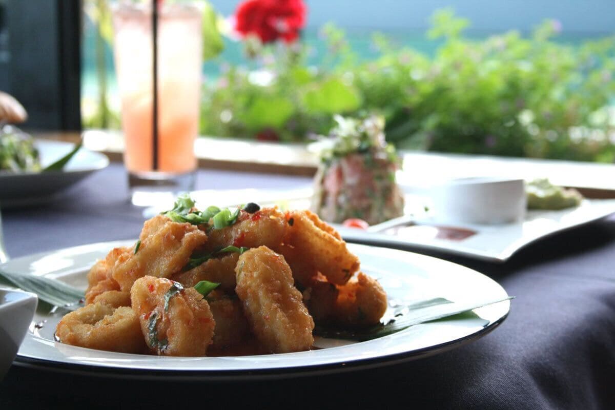 A plate of fried food garnished with herbs, a glass of iced drink, and a view of greenery and a red flower in the background.