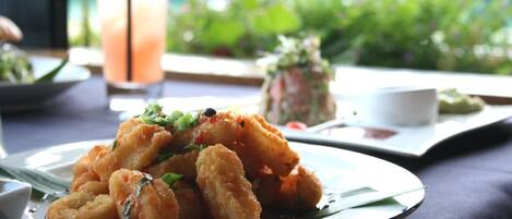 A plate of fried food garnished with herbs, a glass of iced drink, and a view of greenery and a red flower in the background.