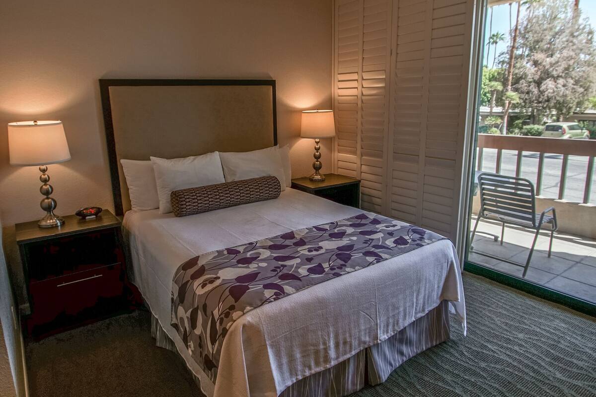 A hotel room with a bed, two bedside lamps, a ceiling fan, and a view of the outdoors through a sliding glass door.