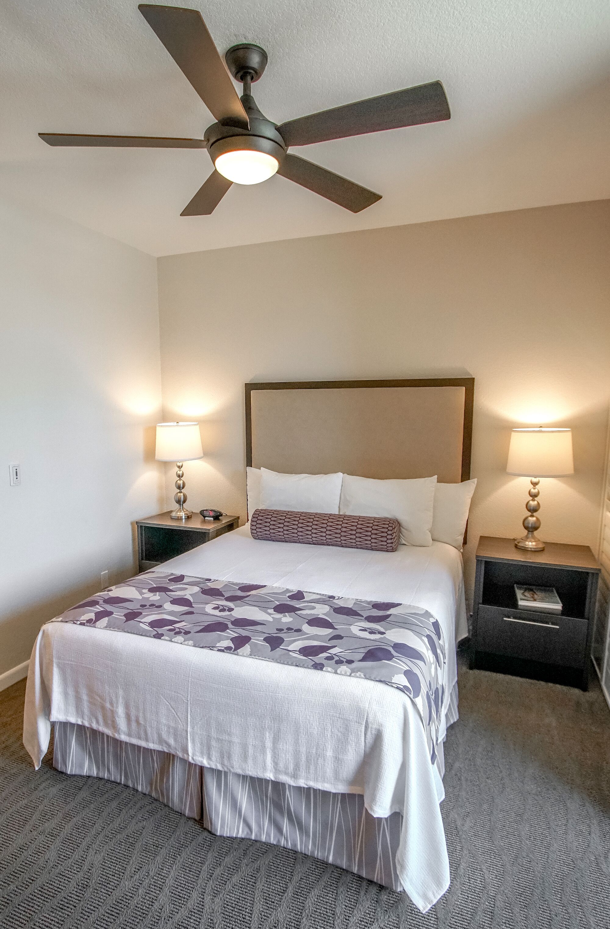 A hotel room with a bed, two bedside tables with lamps, a ceiling fan, and a carpeted floor.