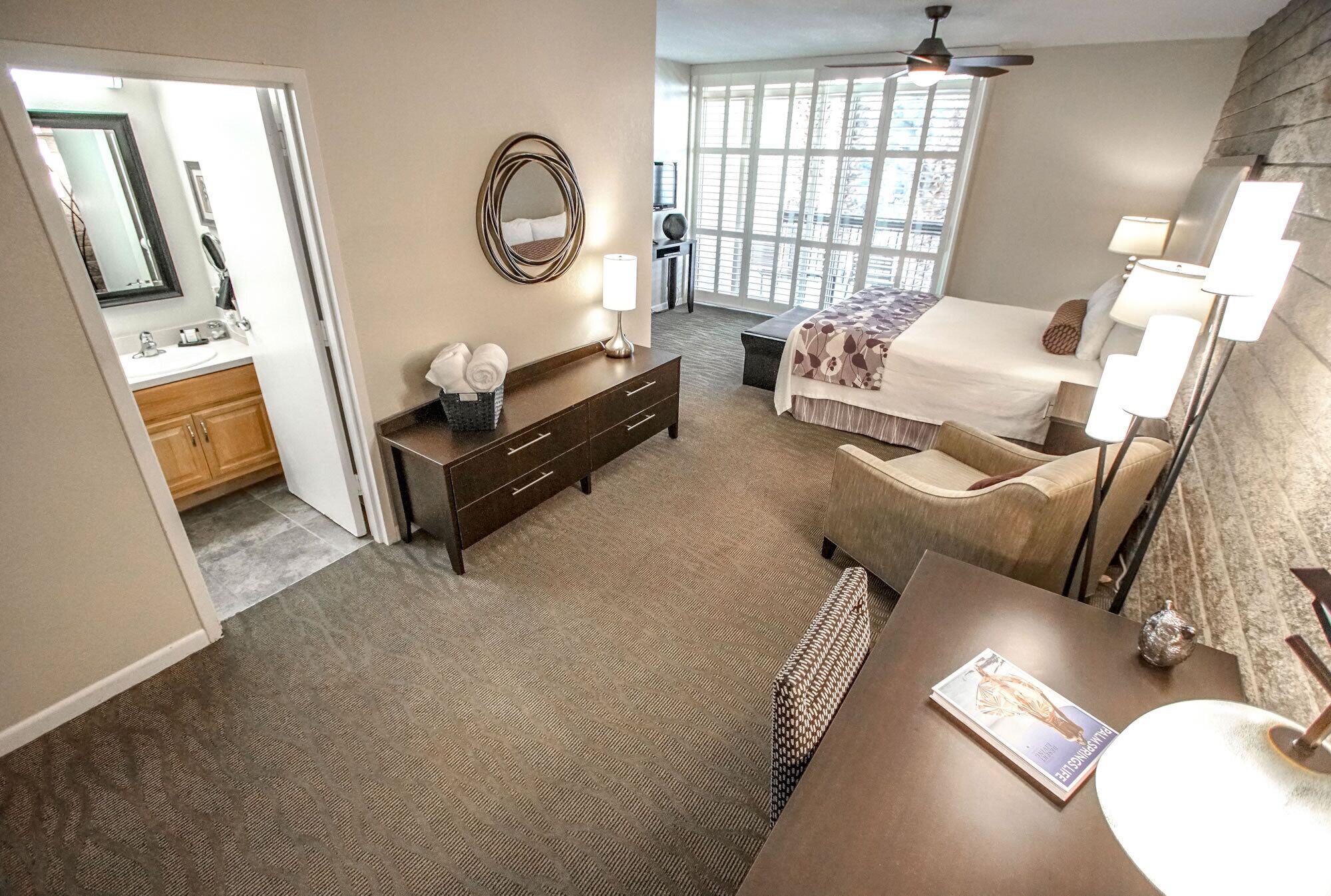 A hotel room with a bed, bedside lamps, a desk with a lamp, a chair, a small table, a mirror, and a window with a view.