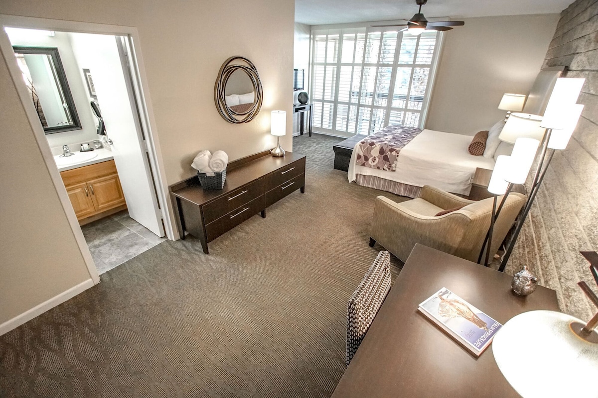 A hotel room with a bed, bedside lamps, a desk with a lamp, a chair, a small table, a mirror, and a window with a view.