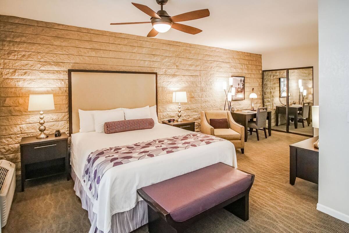 A hotel room with a large bed, a desk, a chair, a bench, a ceiling fan, and a mirror.