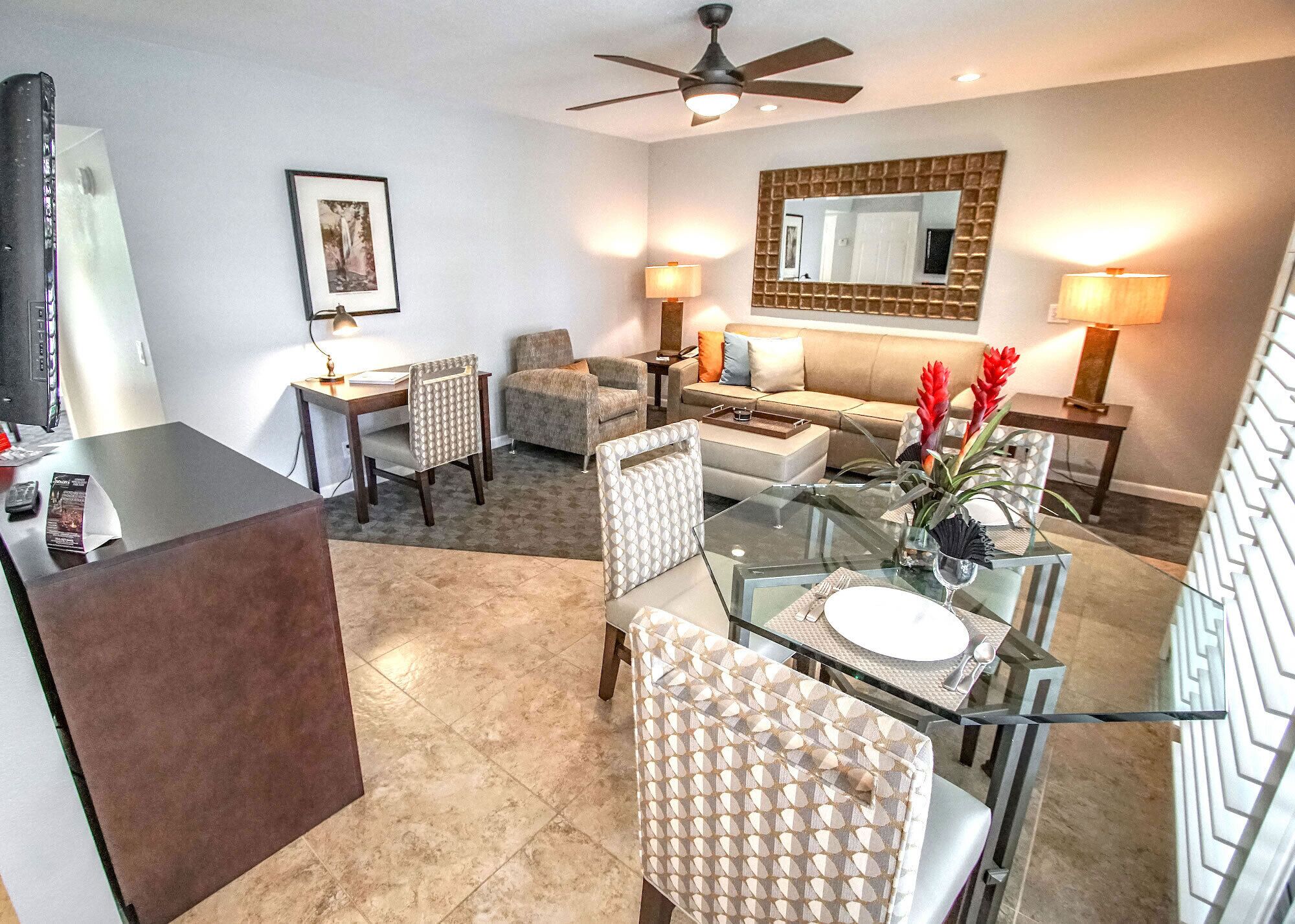A modern living room with a glass dining table, comfortable seating, a sofa, and a large mirror.