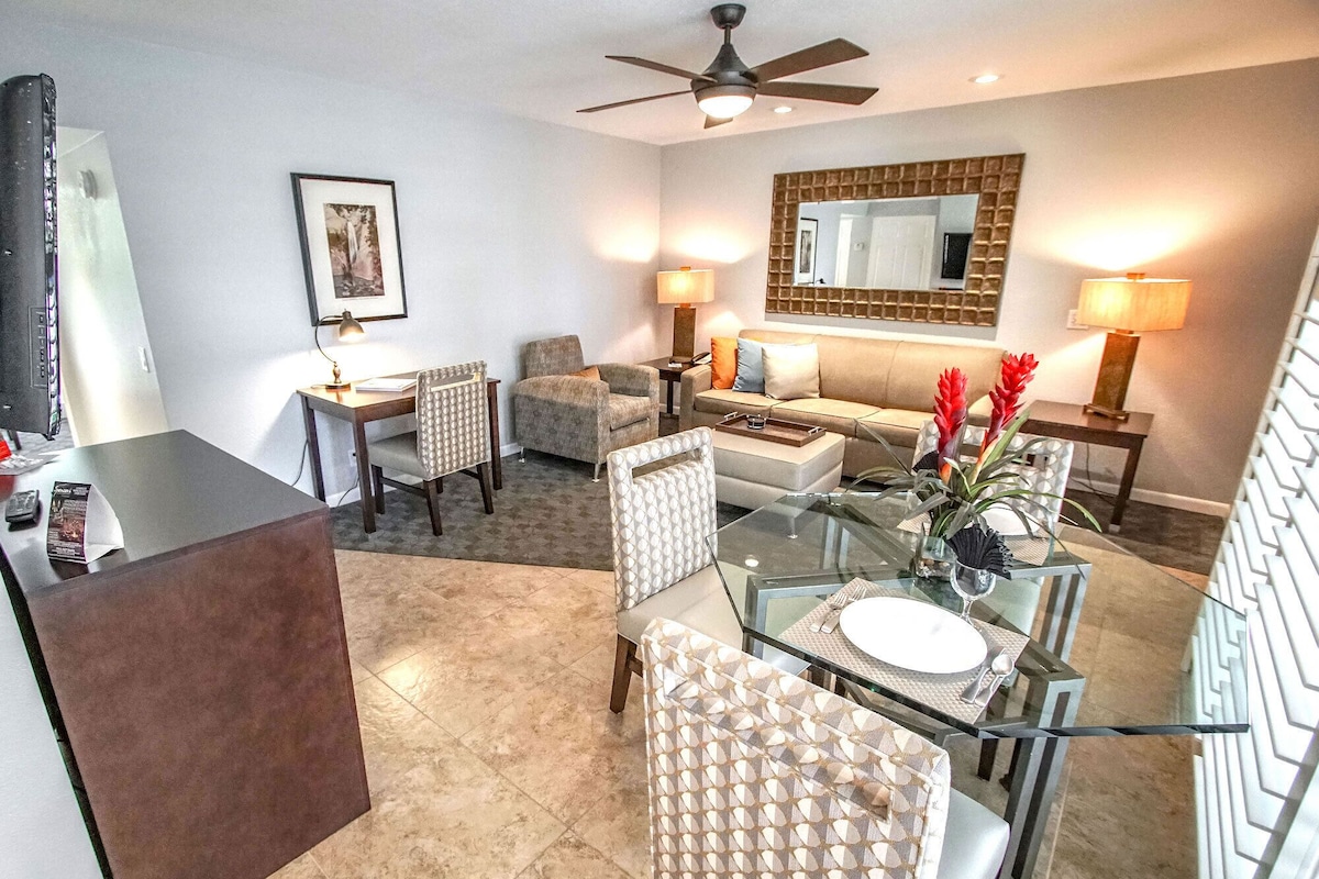 A modern living room with a glass dining table, comfortable seating, a sofa, and a large mirror.