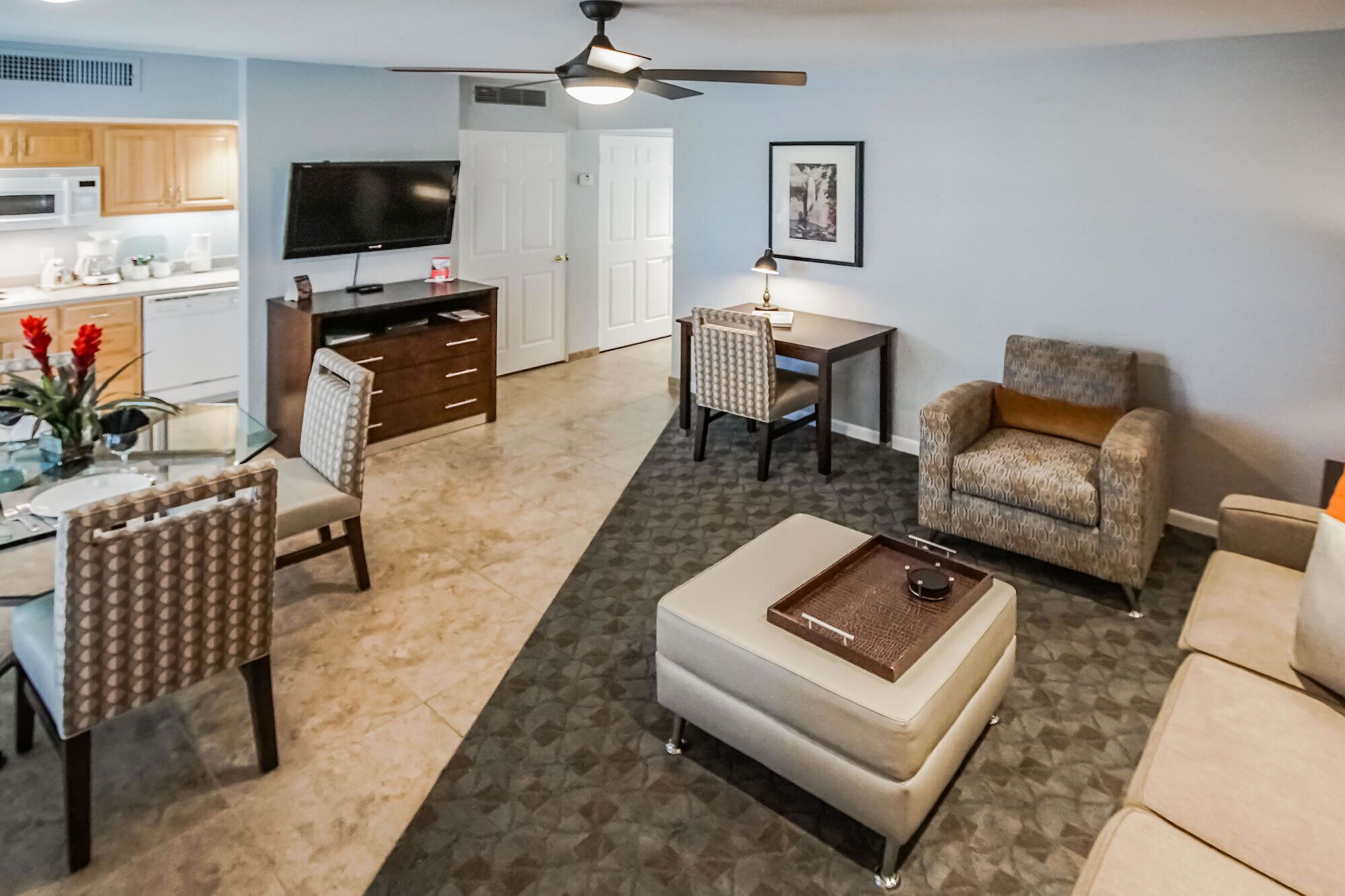 A living room with a sofa, armchair, coffee table, dining table, and chairs. There is a television, a ceiling fan, and a framed picture on the wall.