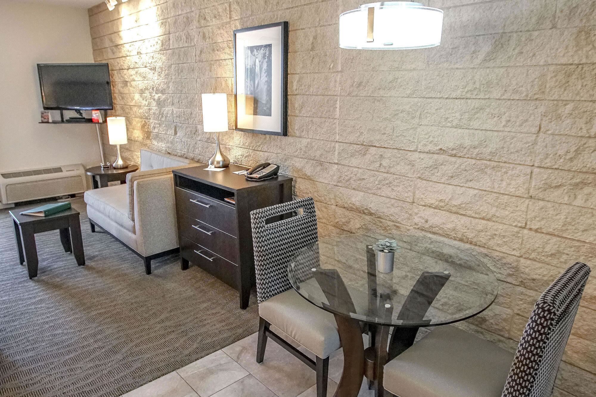 Une chambre d’hôtel avec un mur en pierre, un téléviseur à écran plat, un bureau avec une lampe, une chaise et une table à manger avec des chaises.