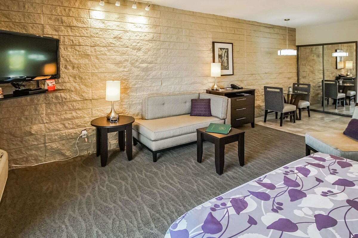 A hotel room with a sofa, a small table with a lamp, a TV, a desk with a chair, and a bed with a floral bedspread.