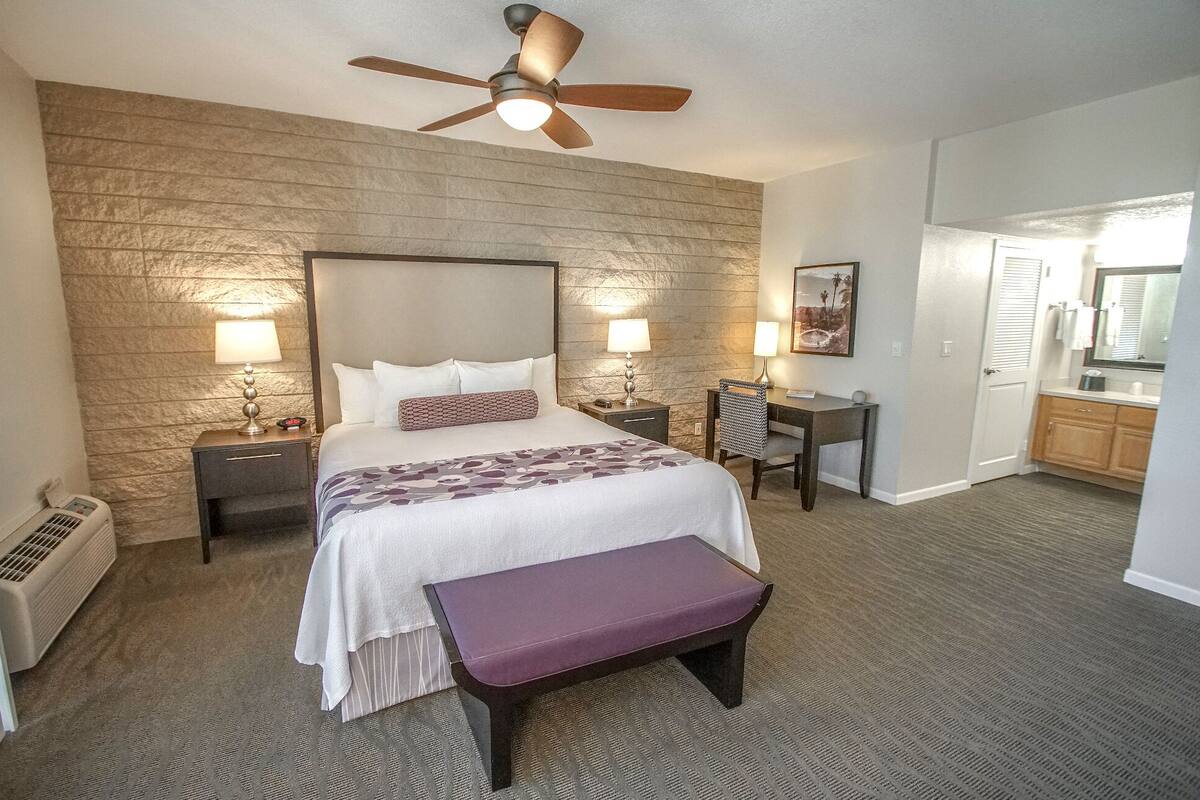 A hotel room with a large bed, two bedside tables with lamps, a ceiling fan, a bench, a desk, and a bathroom visible through an open door.