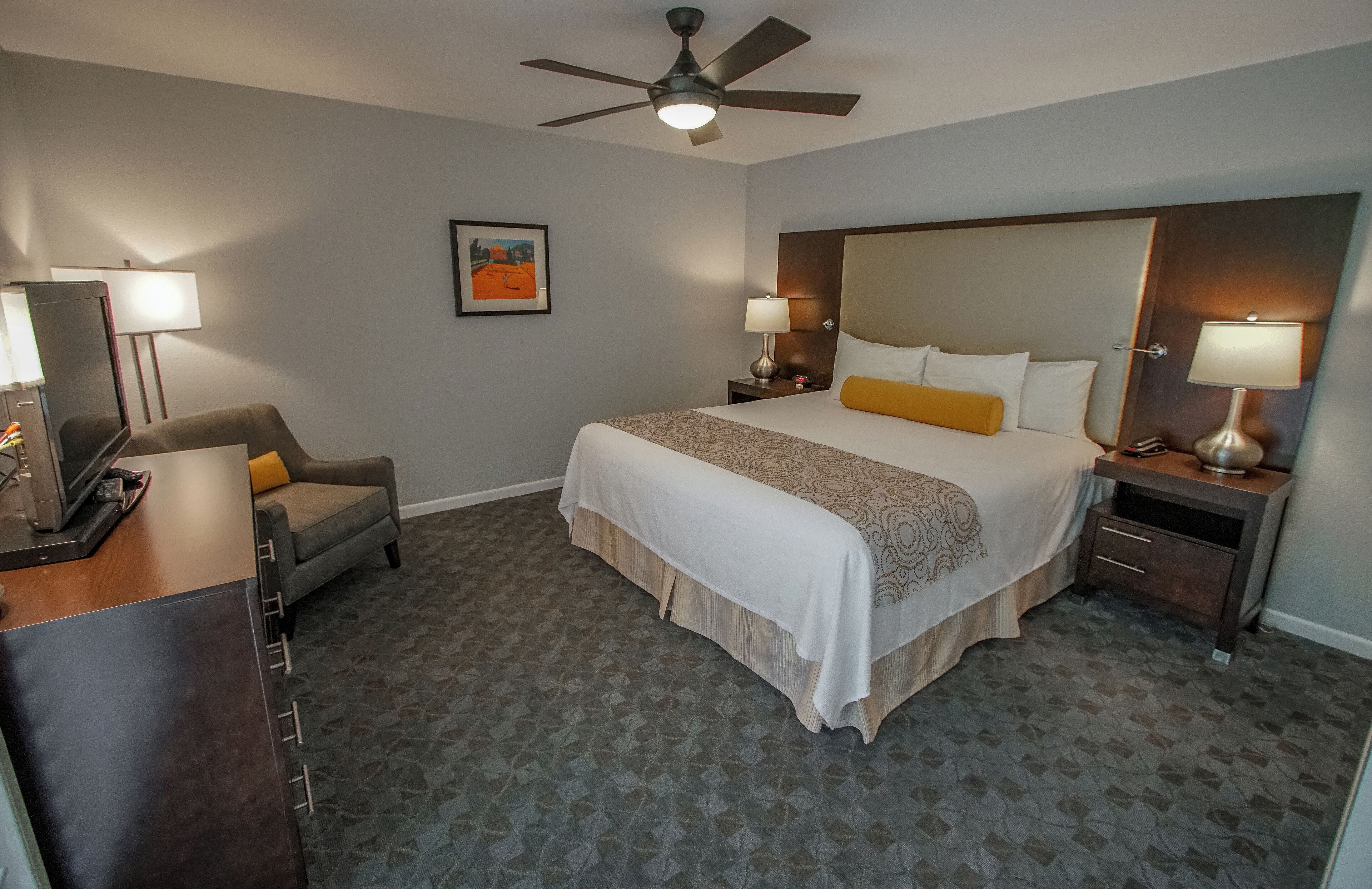 Quarto de hotel com uma cama grande, duas mesas de cabeceira com luminárias, uma escrivaninha com luminária, uma cadeira e um ventilador de teto.
