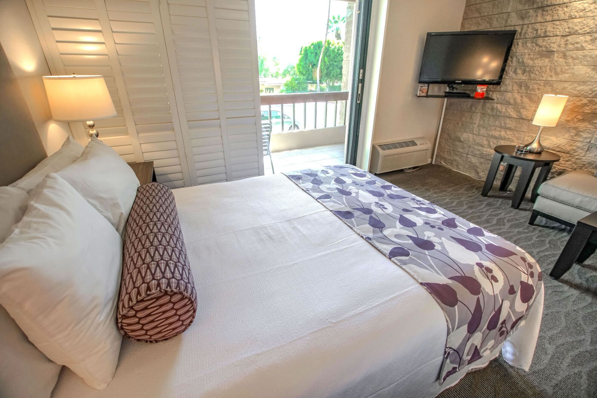 A hotel room with a bed, two bedside lamps, a television on the wall, and a small table with a lamp.