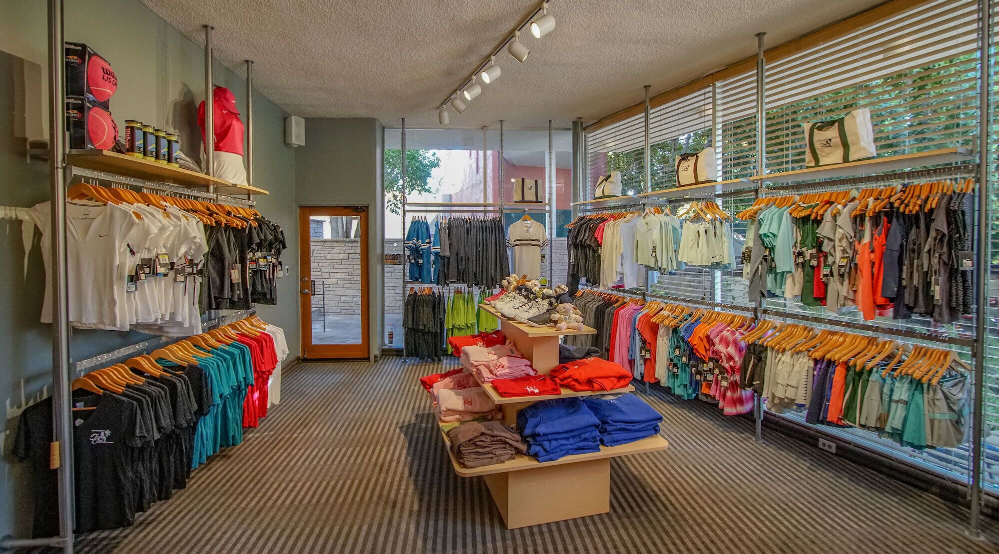 Un magasin de vêtements avec des portants remplis de chemises et de pantalons, une table de présentation où sont pliés des vêtements, et une porte fermée au fond.
