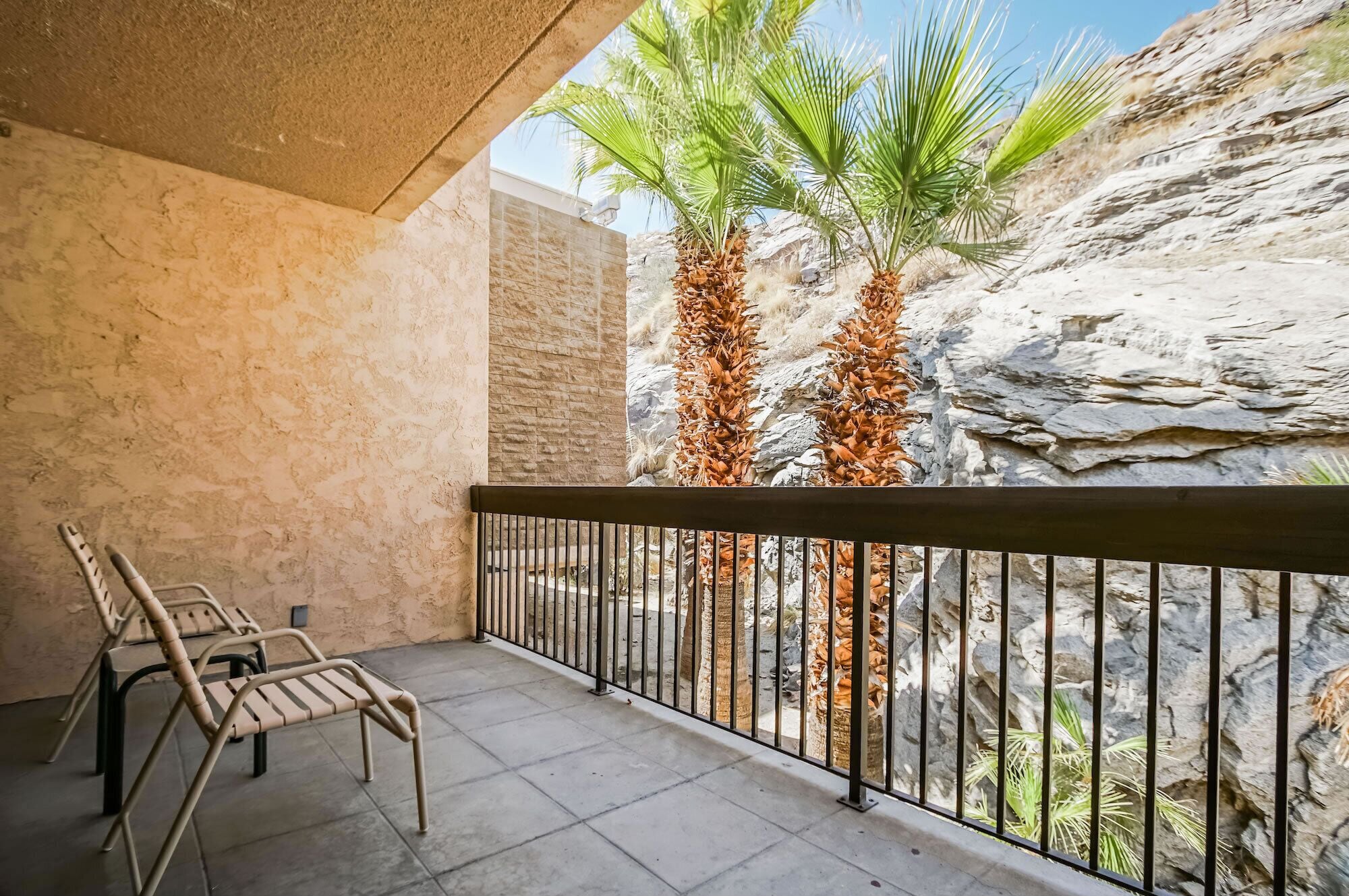 A balcony with a stone wall, two palm trees, and two chairs.