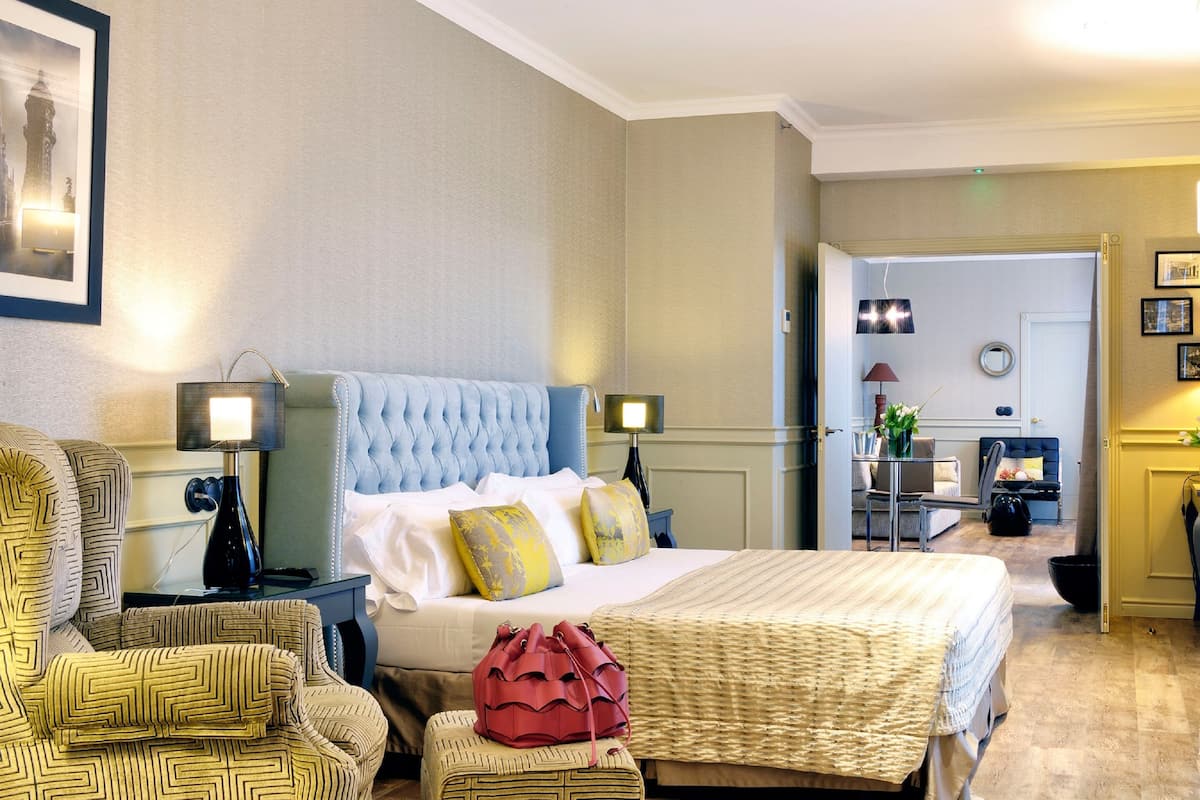 Une chambre d’hôtel avec un grand lit, deux tables de chevet avec des lampes, une chaise, une petite table avec un vase et une porte donnant sur une autre pièce.