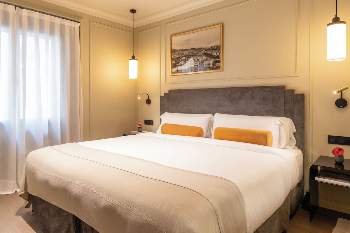 Une chambre d’hôtel avec un grand lit, une table de chevet avec une lampe, un tableau encadré au mur et une fenêtre avec des rideaux.