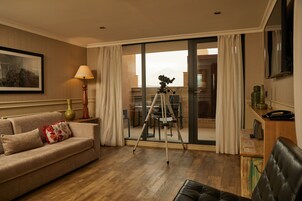 Quarto de hotel com sofá, tripé e área de jantar visível pelas portas de vidro.