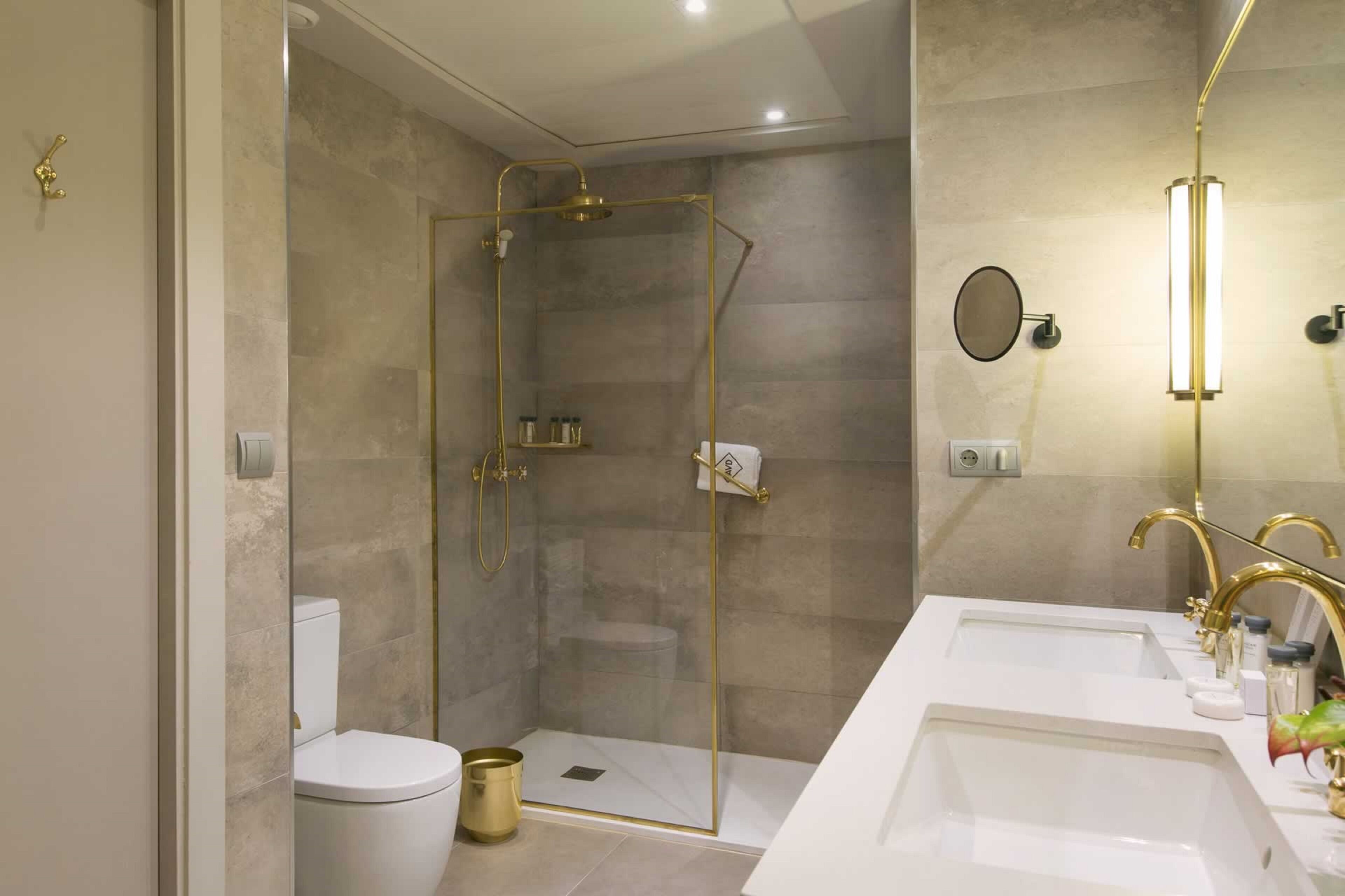 A modern bathroom with a glass-enclosed shower, a white toilet, and a double sink with gold fixtures.