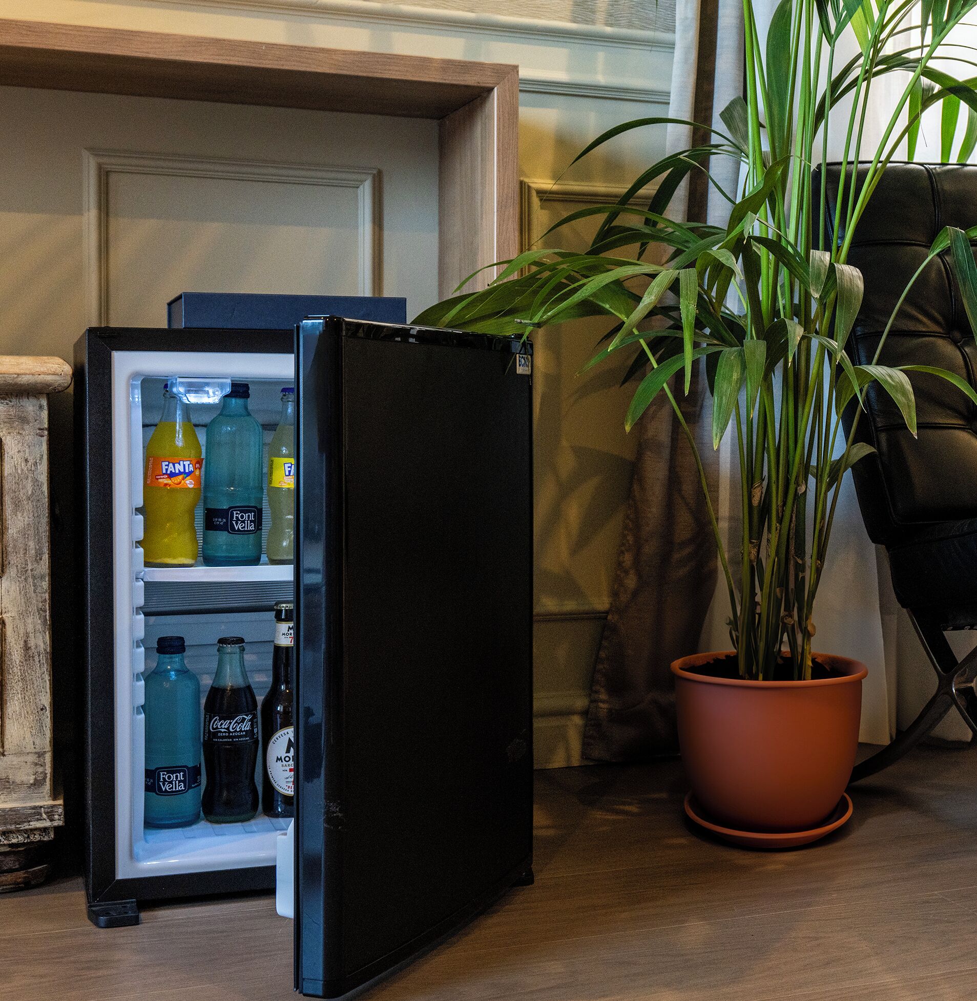 A refrigerator with beverages inside, a potted plant beside it, and a chair in the background.