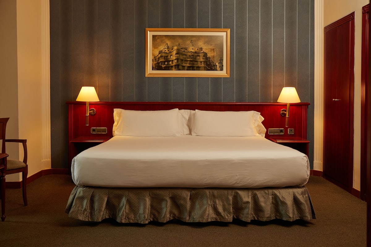 Un lit bien fait, avec du linge de lit blanc, une tête de lit en bois et un pied de lit orné d’une jupe. Deux lampes de chevet sont placées de chaque côté du lit, et un tableau encadré est accroché au-dessus.