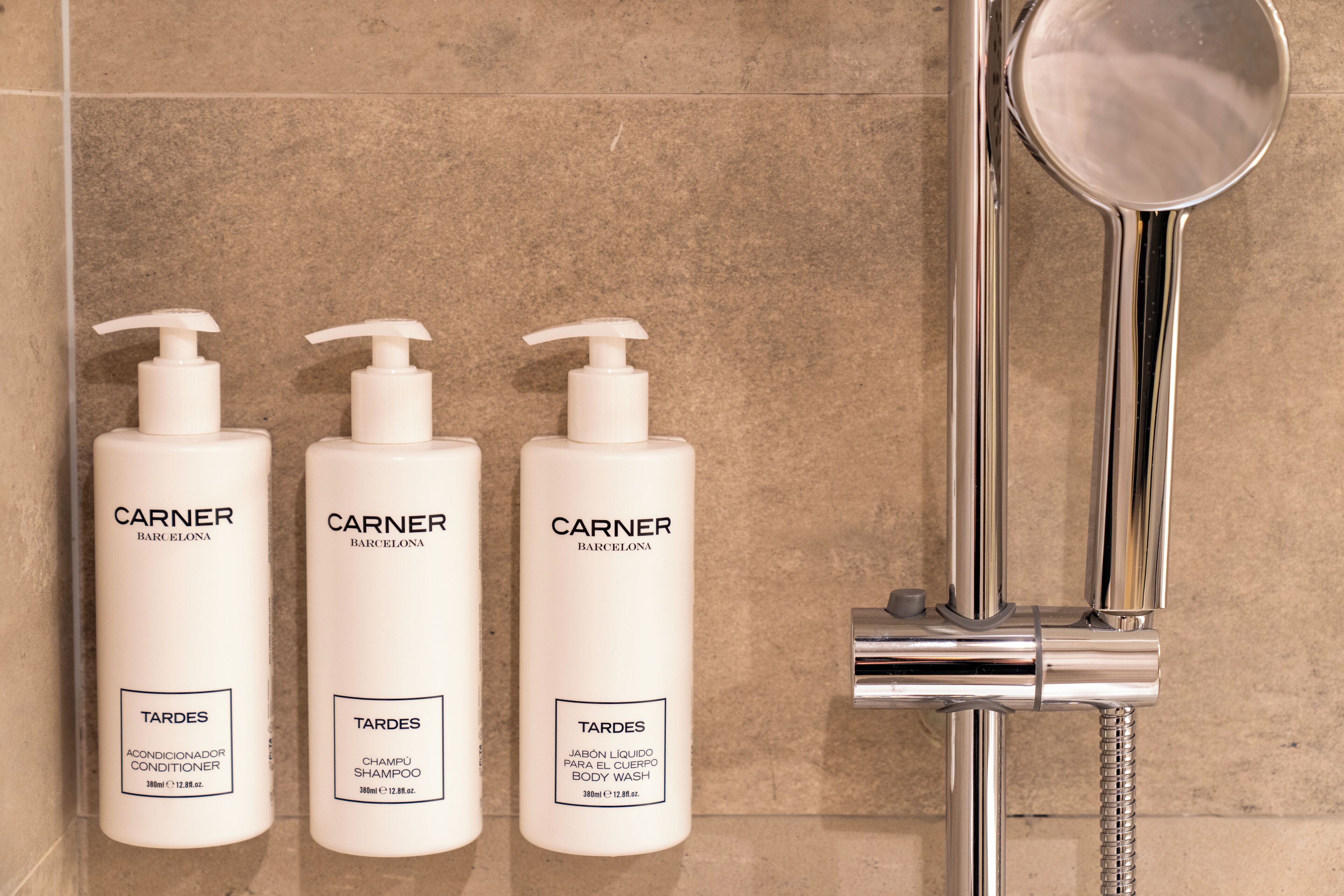 Tres botellas de productos Carner Barcelona: acondicionador, champú y gel de ducha, ubicadas en una pared de azulejos junto a una grifería de ducha cromada.