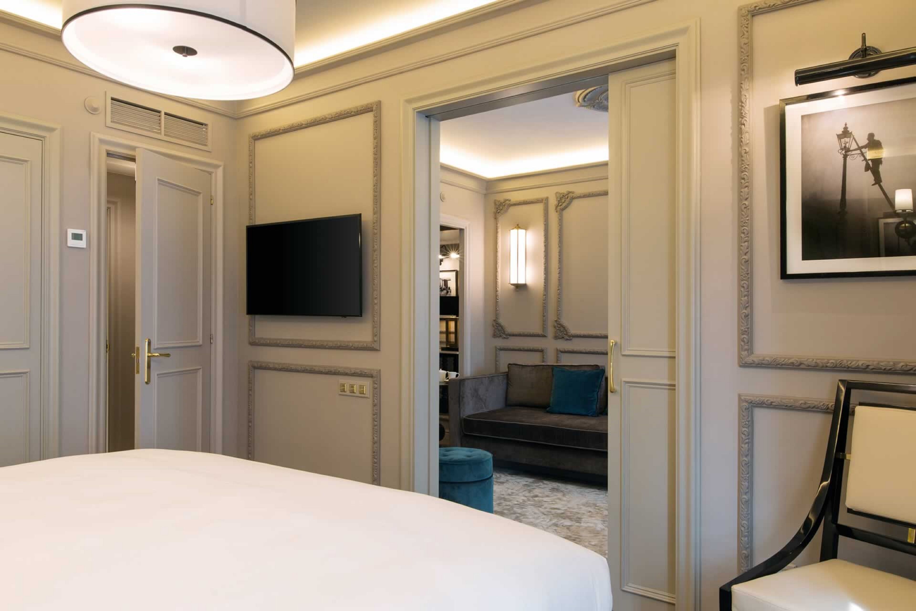 A hotel room with a large bed, a flat-screen TV mounted on the wall, a sofa in the adjacent room, and a framed picture on the wall.
