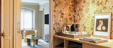 A hotel room with a wooden desk, a coffee maker, a kettle, a framed picture, and a bookshelf with a book titled "LUXURY HOTELS".