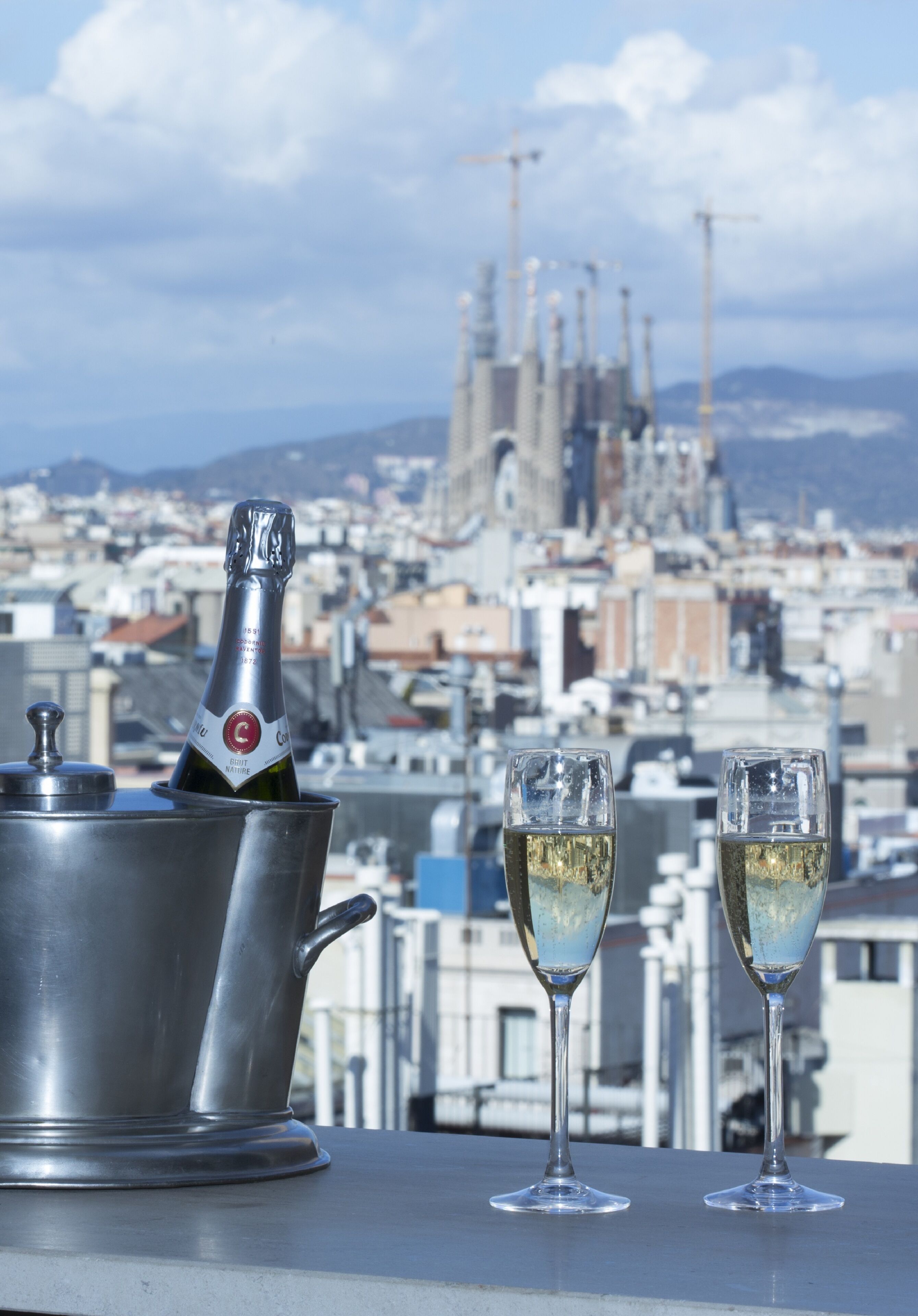 A champagne bottle in a metal bucket with two glasses on a rooftop, overlooking a cityscape with buildings and a cathedral.