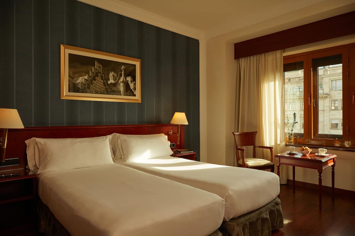 Une chambre d’hôtel avec deux lits, une tête de lit en bois, un tableau encadré au mur, une fenêtre avec des rideaux, une petite table sur laquelle se trouve un saladier rempli de fruits et une chaise.