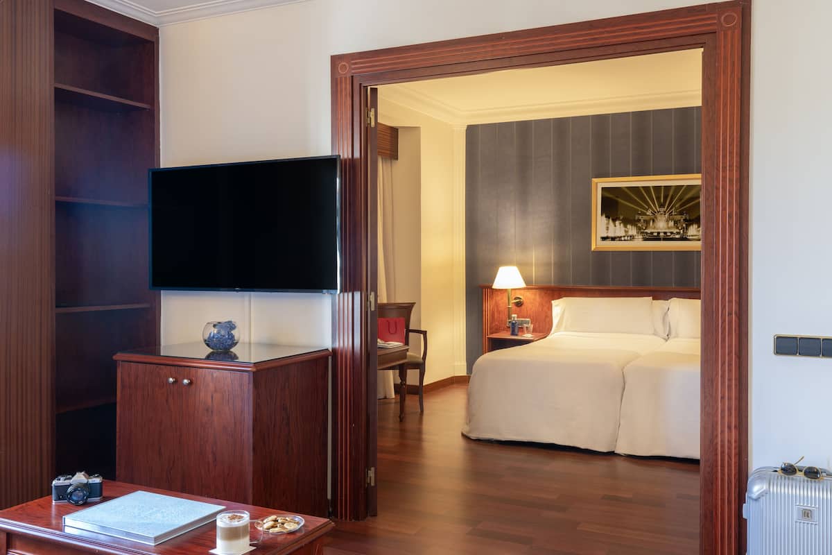 Une chambre d’hôtel avec un bureau en bois, un téléviseur à écran plat, un lit, une table de chevet avec une lampe et un tableau encadré au mur.