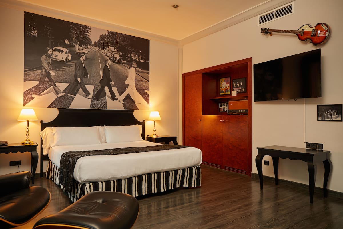 Une chambre d’hôtel avec un grand lit, deux lampes de chevet, un téléviseur à écran plat, une guitare accrochée au mur et une affiche d’un groupe de musique célèbre.