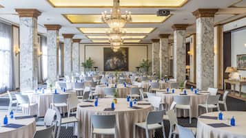 Uma sala de banquetes com mesas redondas, cadeiras brancas, lustres e colunas de mármore.